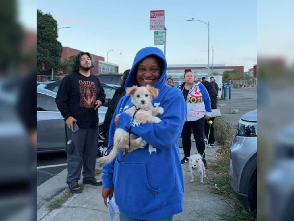 A woman in a blue hoodie holds a small white dog on a sidewalk, with several people, parked cars, and a street sign in the background.