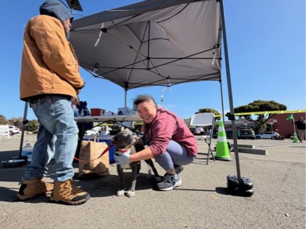 A woman crouches beside a dog under a pop-up canopy in a parking lot, with another person standing nearby and table items in the background.