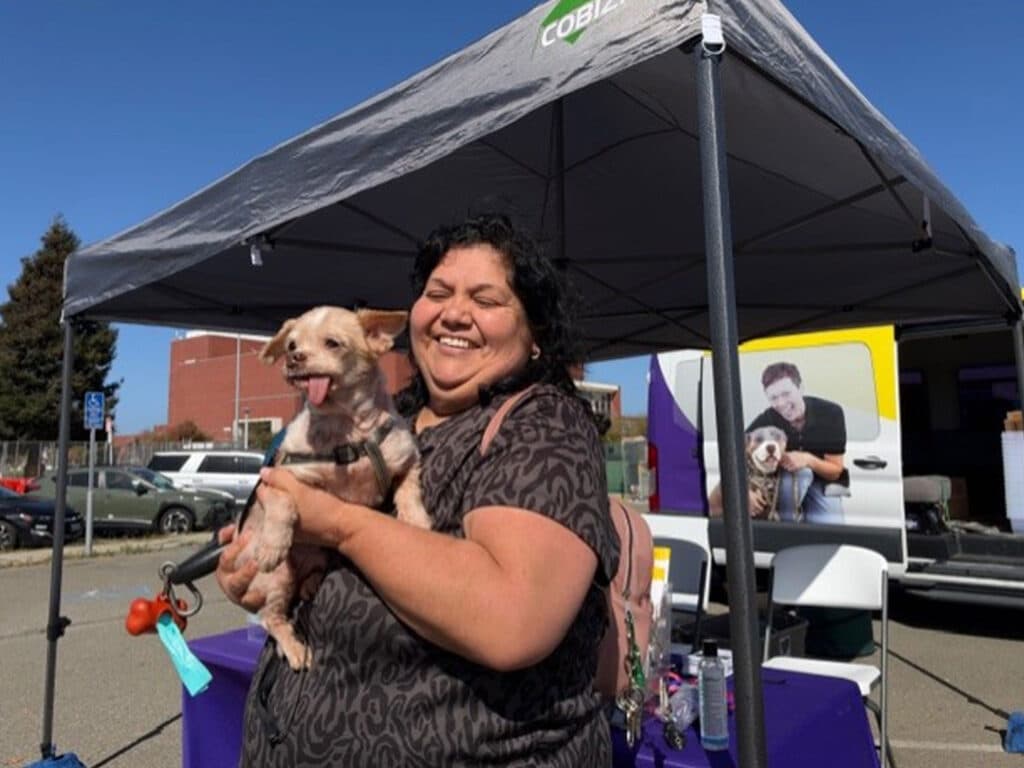 A woman holding a small dog stands under a gray canopy tent at an outdoor event, with tables, chairs, and a van in the background.