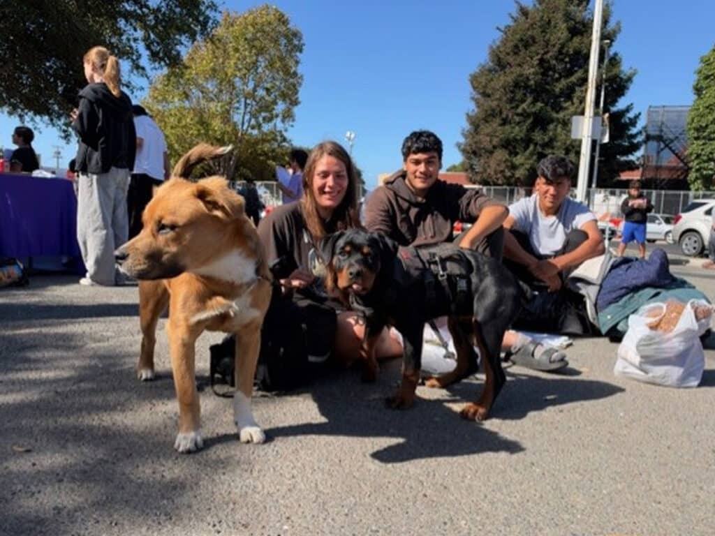 Three young people crouch beside two leashed dogs in a parking lot, with a food drive or community event visible in the background.