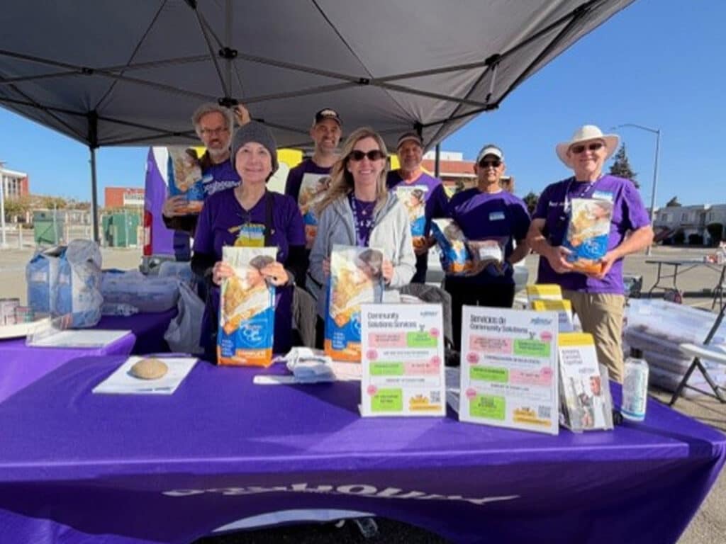 A group of volunteers in purple shirts stands behind a table with flyers and packaged food under a canopy at an outdoor vaccine clinic and food pantry event.