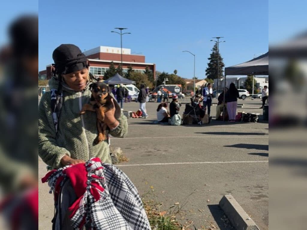 A woman in a black hat holds a small dog and a bundle of blankets in a parking lot, with tents, people, and a building in the background.
