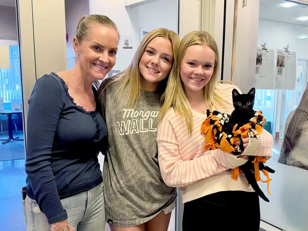 Three women pose in an indoor shelter lobby, with the woman on the right holding a small black cat wrapped in an orange-and-black blanket.