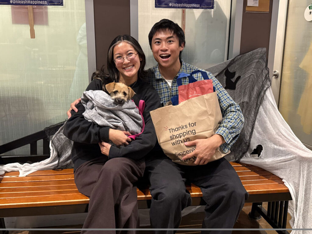 A smiling couple sits on a bench holding a small dog wrapped in a blanket, with a shopping bag beside them in an indoor adoption setting.