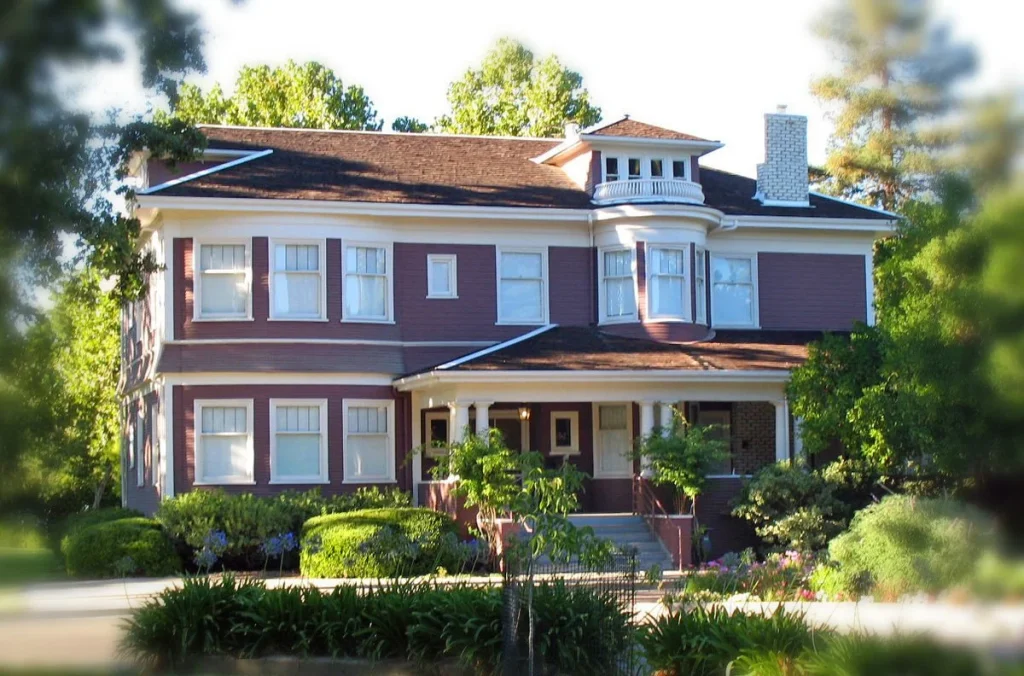 A large two-story house with a brown roof, white trim, and a front porch sits behind trimmed shrubs and trees.
