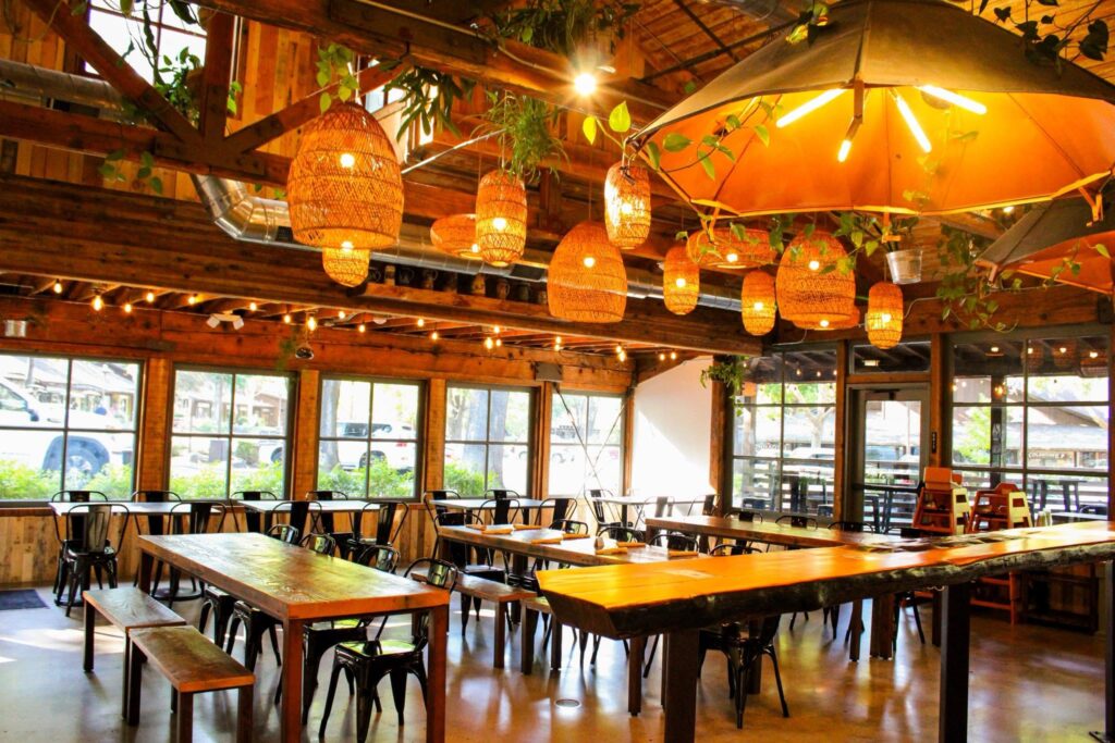 A rustic brewery dining room with wooden tables, benches, and hanging woven lanterns under exposed beams, with large windows and an orange patio umbrella.