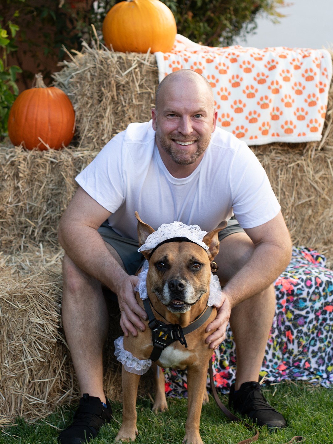 A bald man in a white T-shirt crouches behind a brown dog wearing a white bonnet, with hay bales, pumpkins, and patterned blankets in the background.