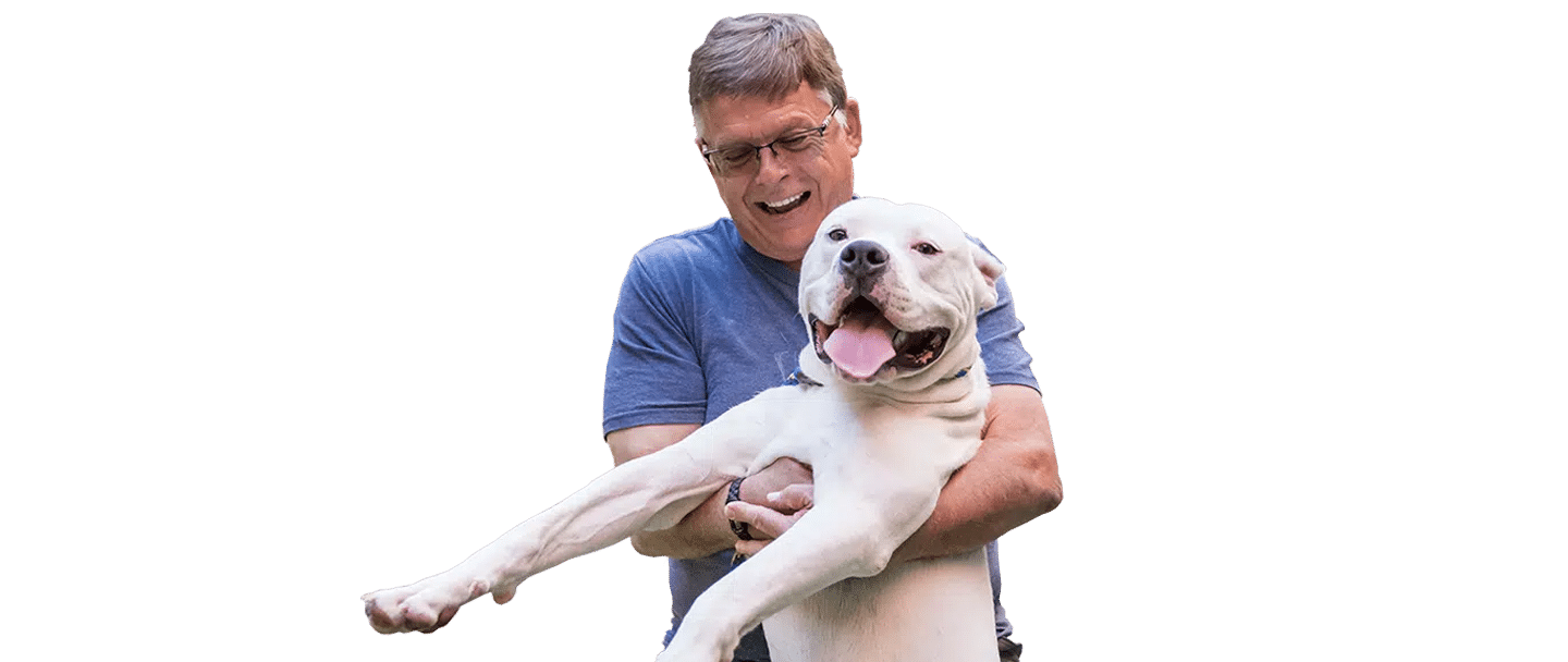 A man in a blue shirt holds a white dog, Joybound, with the dog’s front legs extended against a transparent background.