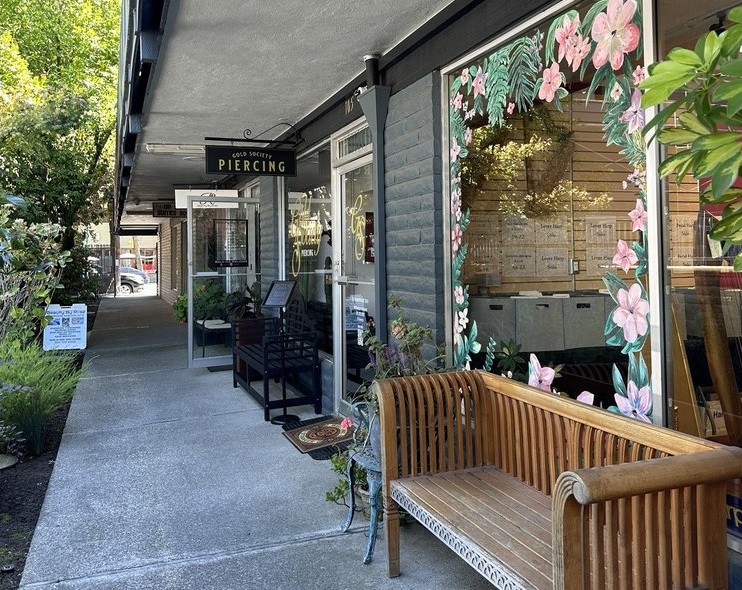 The storefront of Gold Society Piercing with a covered sidewalk, glass windows, and a wooden bench decorated with pink floral window trim.