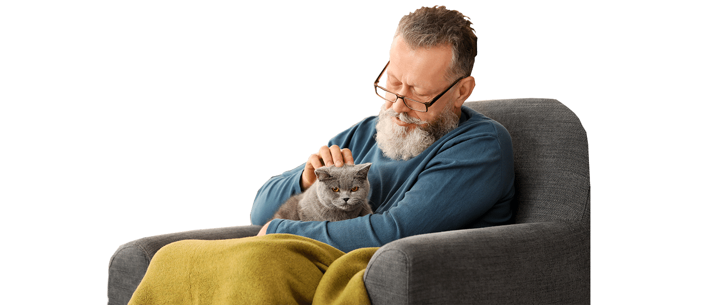 A man with a gray beard and glasses sits in a gray armchair, gently holding a gray cat under a yellow blanket, with Joybound included.