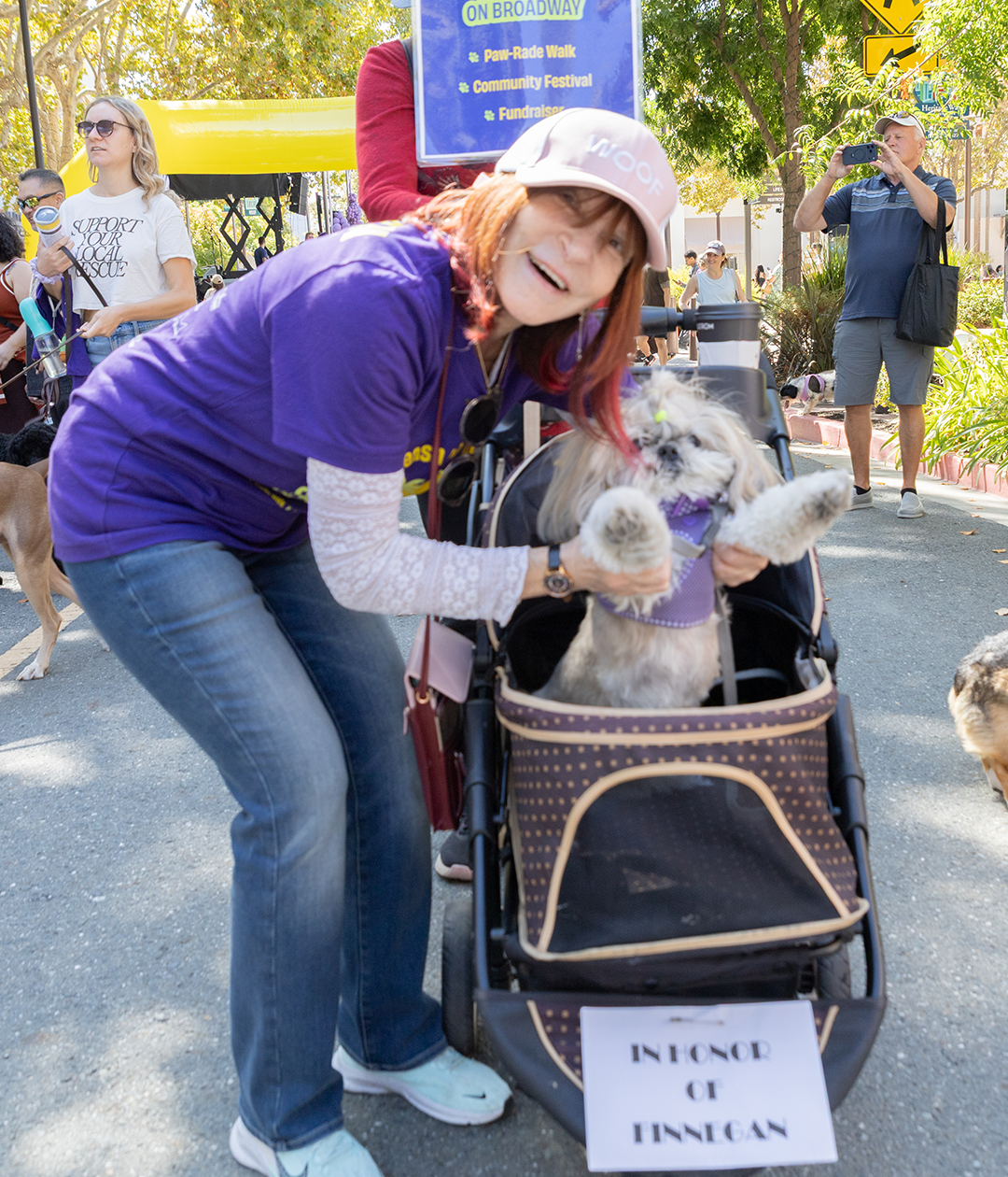 A woman in a purple shirt and pink cap poses with a small dog in a stroller, with a sign reading "IN-N-OUT OF FINNIGAN" at a street festival.