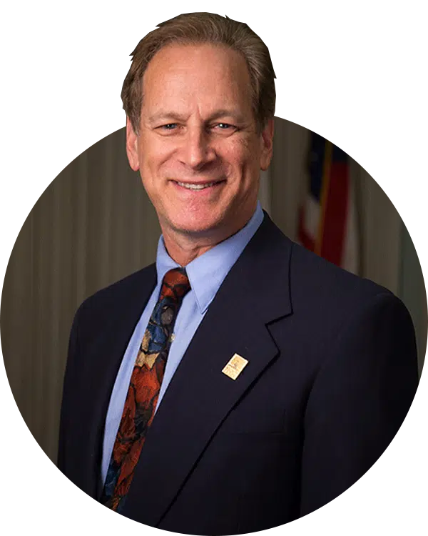A man in a dark suit, blue shirt, and patterned tie poses for a portrait with a small lapel pin against a blurred indoor background.
