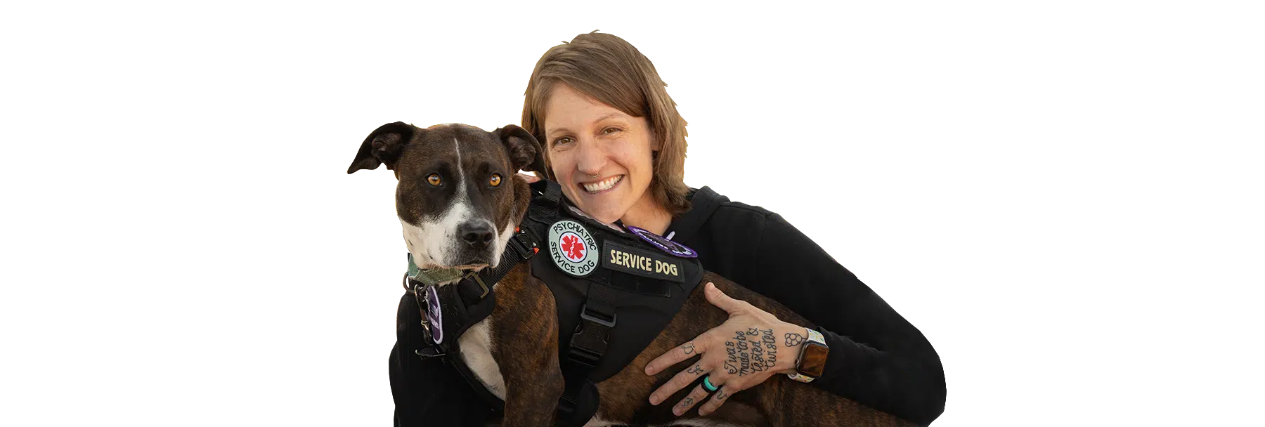 A woman embraces a brown-and-black Psychiatric Service Dog wearing a service vest against a black background.