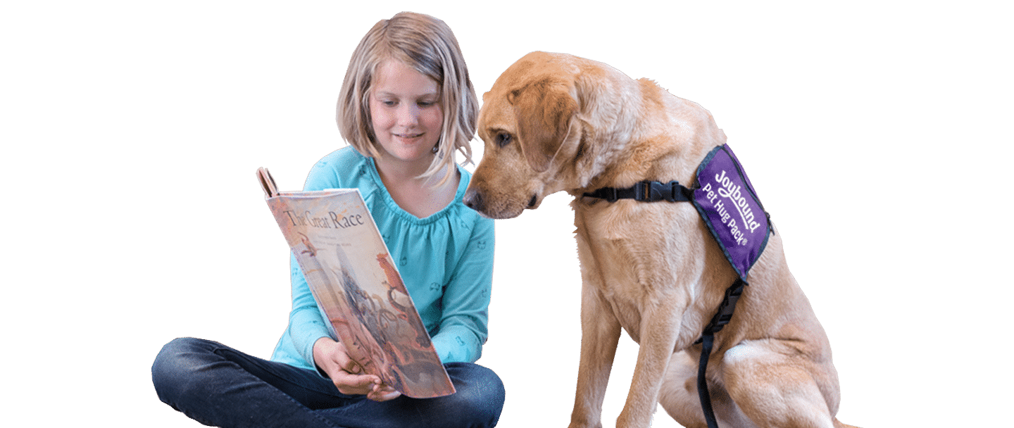A young girl sits beside a yellow Labrador wearing a purple Joybound vest while holding an open book, against a white background.