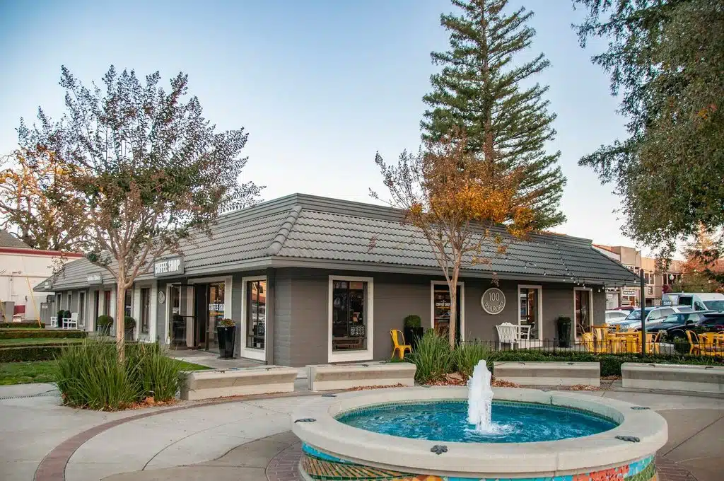A low, single-story building with large windows sits behind a small circular fountain and landscaped courtyard, with tall trees and parked cars in the background.