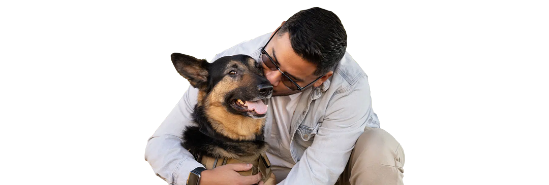 A man kneels and hugs a Service Dog, a German shepherd wearing a tan harness, against a white background.