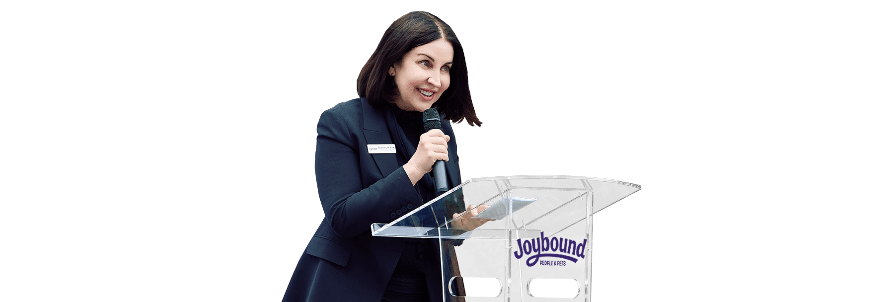 A woman speaking at a clear podium with a Joybound sign, wearing a dark blazer against a white background.