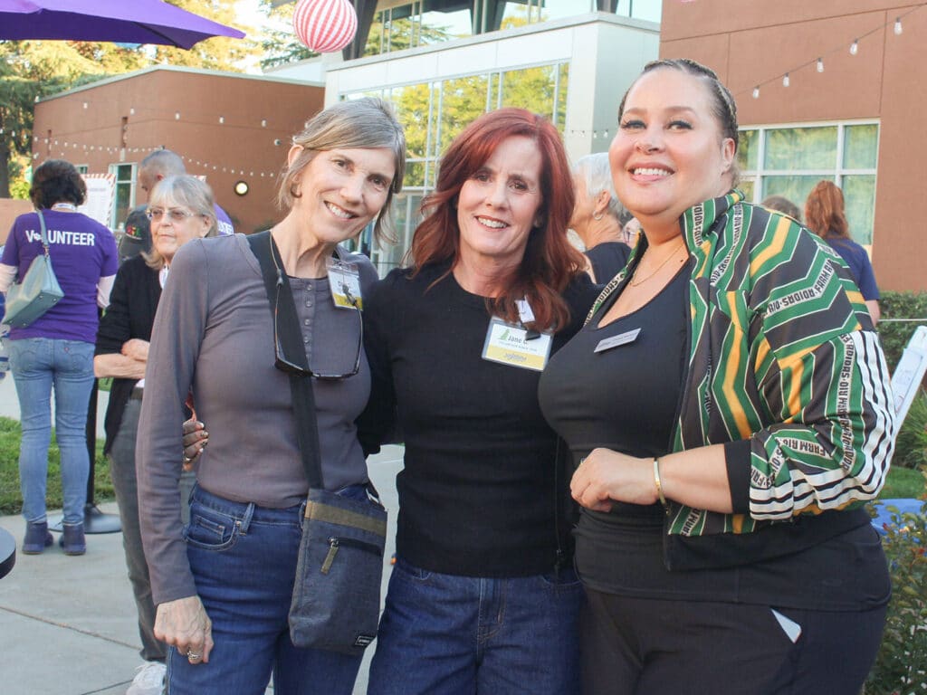 Three women stand together outdoors at a volunteer appreciation dinner, with string lights, patio umbrellas, and other attendees in the background.