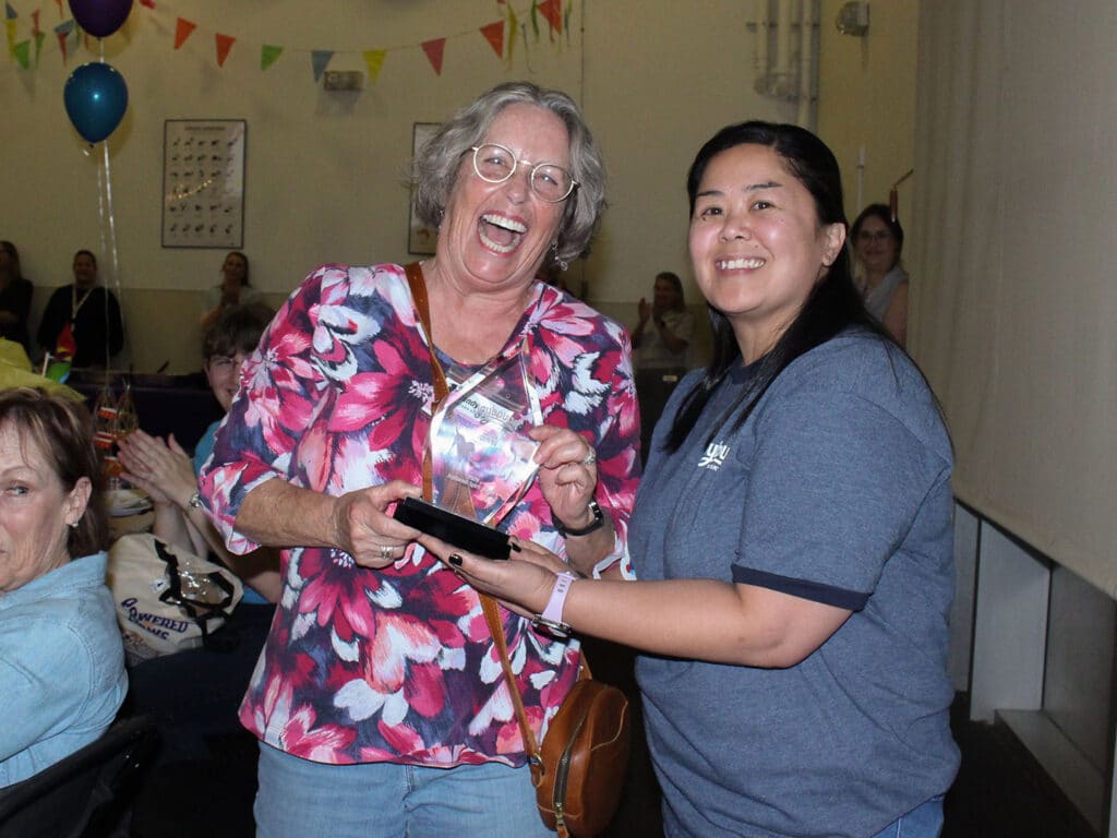 Two women stand in a decorated room as one presents a trophy to the other, with seated attendees and balloons in the background.