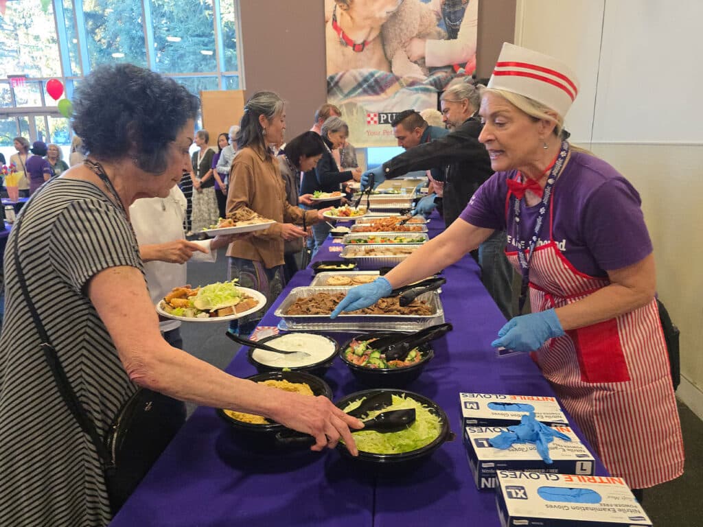 A woman in a red-and-white apron serves food from buffet trays across a purple-covered table, while guests line up with plates in a bright hall.