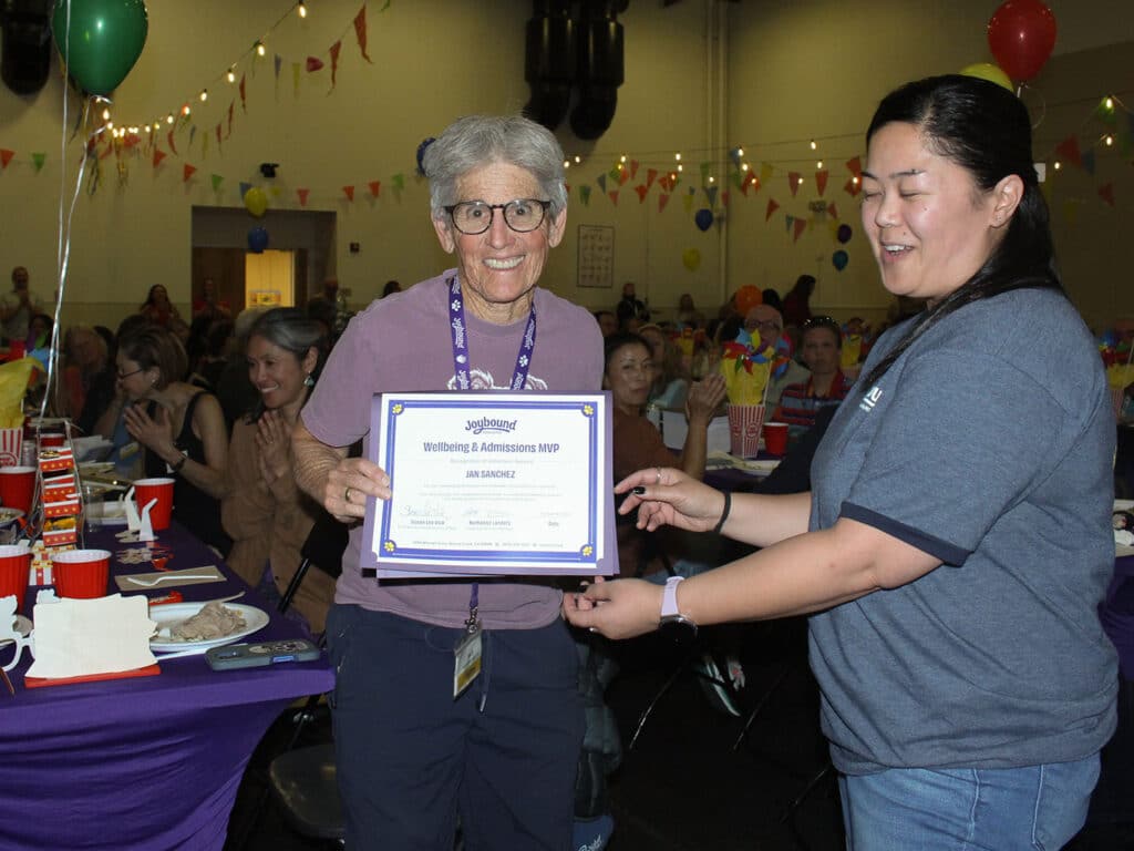 An older woman with glasses holds a framed certificate while a woman in a gray shirt stands beside her at a decorated volunteer appreciation dinner, with seated guests and tables in the background.