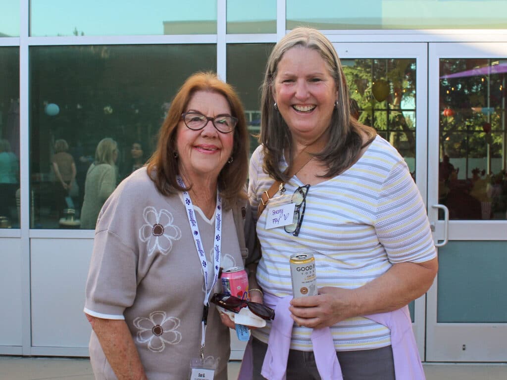Two women wearing lanyards stand together outside a glass entrance, with drink cans in hand.