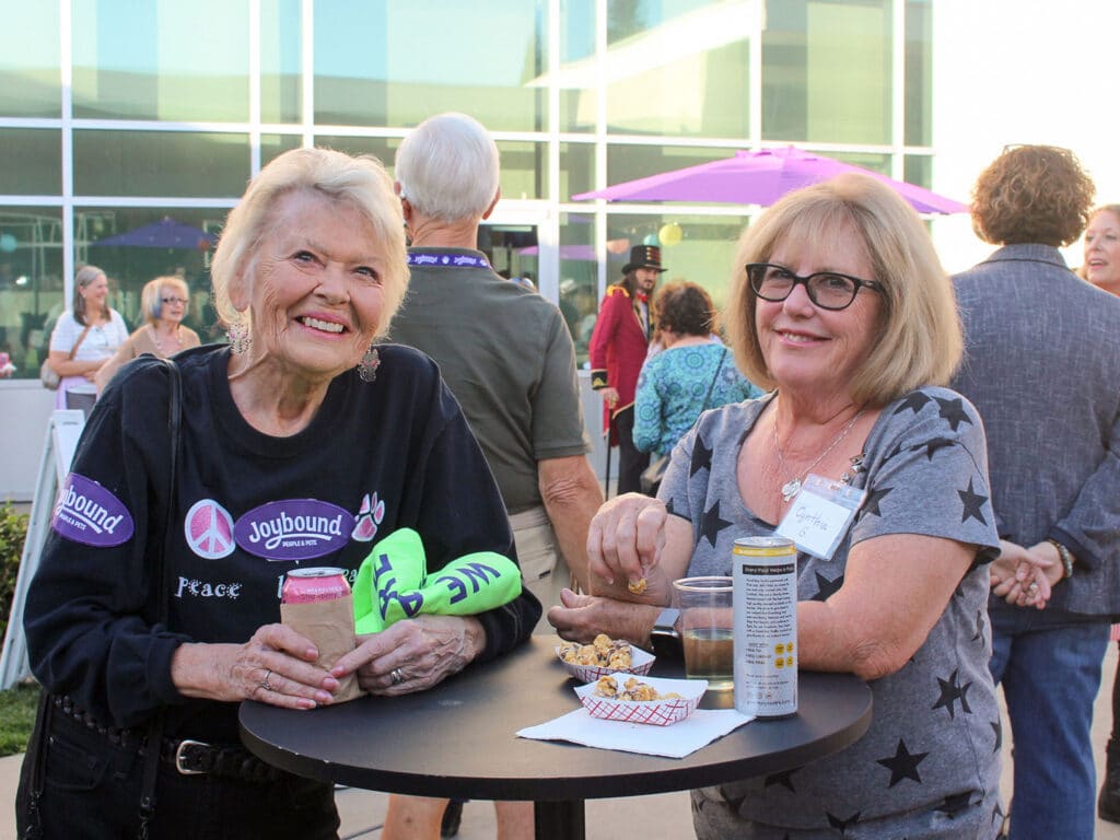Two women stand at a cocktail table during an outdoor volunteer appreciation dinner, with drinks and snacks in front of a glass building.