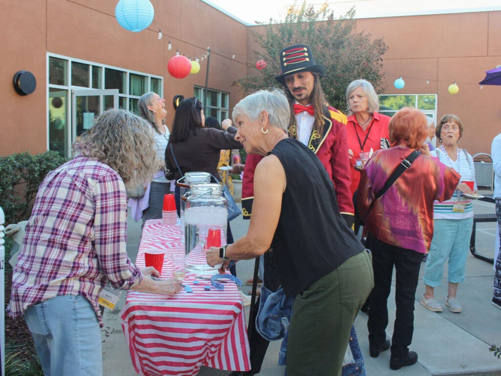 Volunteers and guests stand around a striped serving table at an outdoor dinner, with paper lanterns hanging above and a man in a decorative costume near the center.
