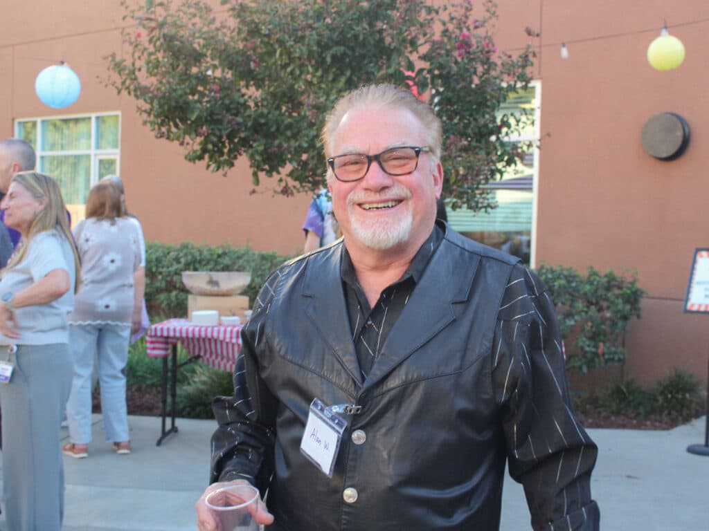 A smiling older man wearing glasses and a black leather jacket stands outdoors holding a plastic cup, with people and a building decorated with hanging lanterns behind him.