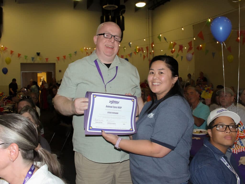 A man and a woman stand together indoors while holding a framed certificate, with seated attendees and balloons in the background.