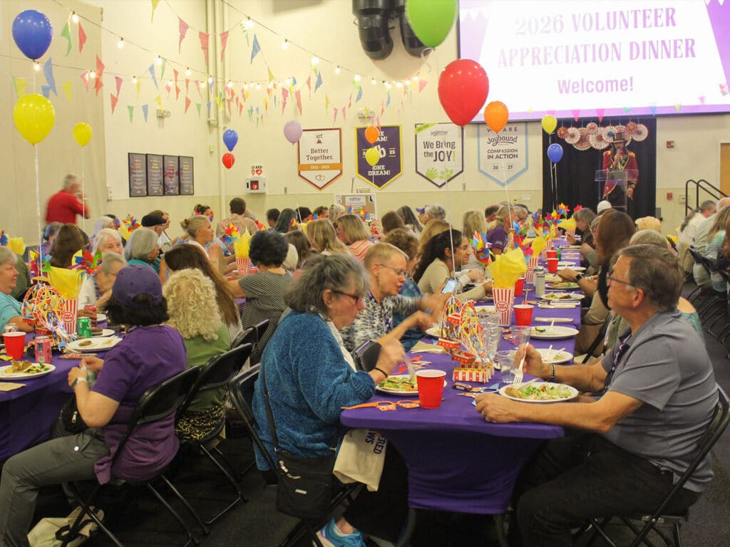Volunteers seated at round tables with purple tablecloths during an appreciation dinner, with balloons, pennant banners, and a welcome screen in the background.