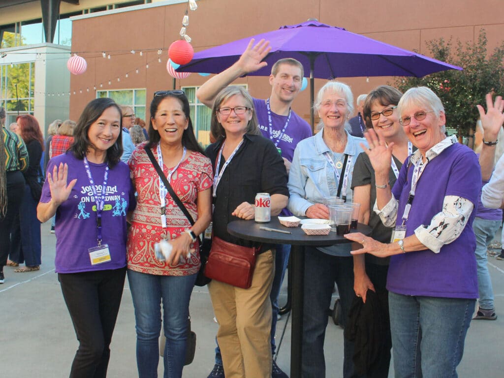 A group of seven volunteers stands around a tall black table outdoors, with several wearing purple shirts and lanyards beneath a purple umbrella.