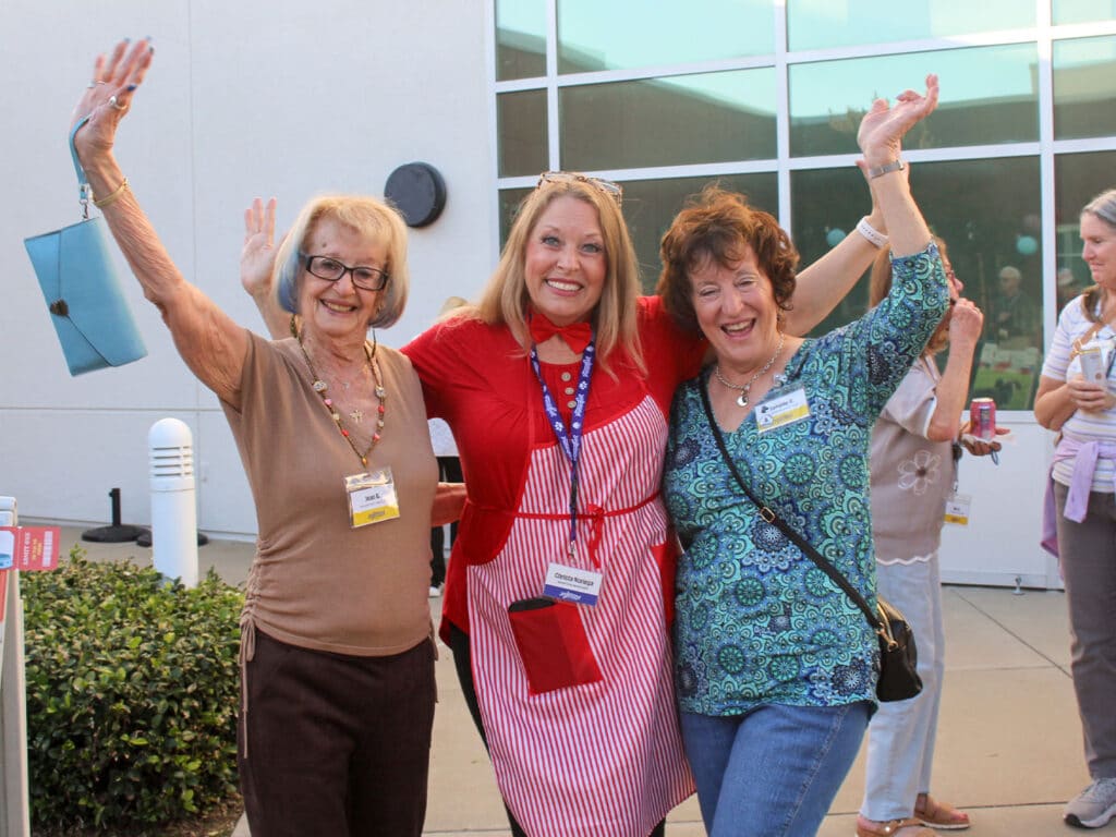 Three women standing together outside a building, two with raised arms and one in a red striped apron, with windows and other attendees in the background.