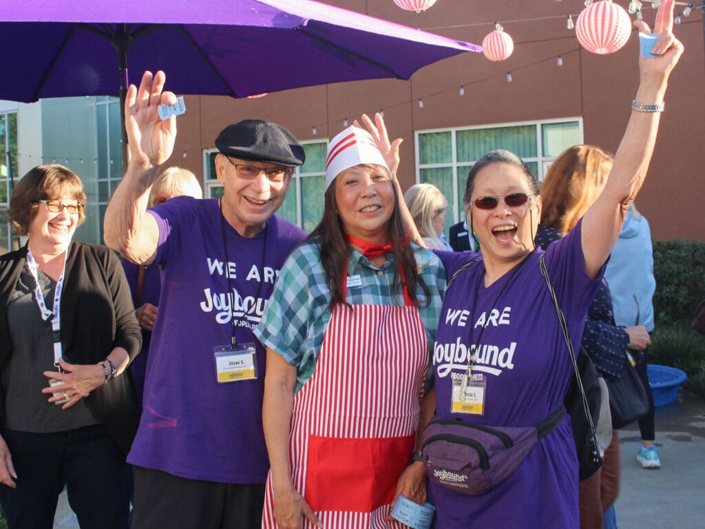 Three volunteers in purple We Are Joybound shirts stand under a purple umbrella, with two raising their arms and one wearing a red-and-white apron and hat, in front of a building with hanging lanterns.