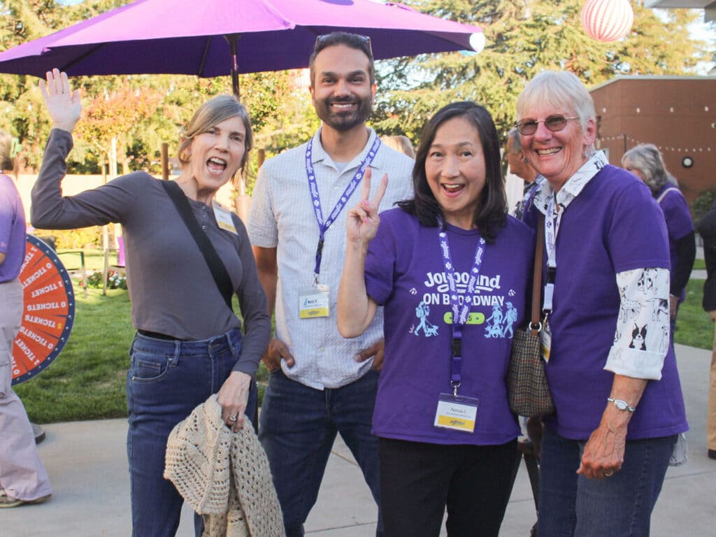Four volunteers standing outdoors under a purple umbrella, with three wearing lanyards and purple shirts, at what appears to be an appreciation event.