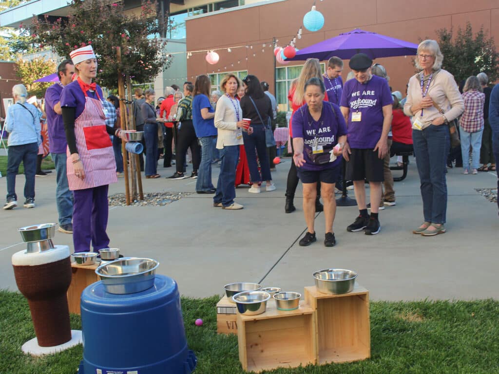 A group of adults stands outdoors at a volunteer appreciation dinner, with a costumed server beside pots and trays on the grass and purple patio umbrellas in the background.