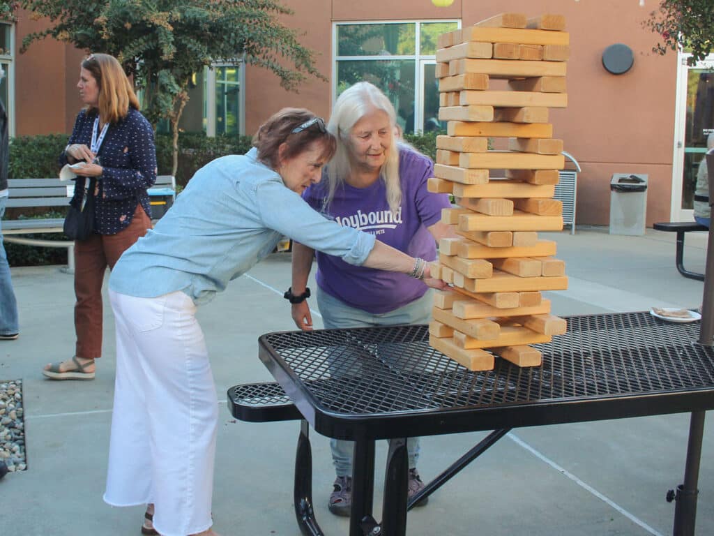 Two women play giant Jenga on an outdoor table, with one leaning in to steady the wooden block tower beside a courtyard building.
