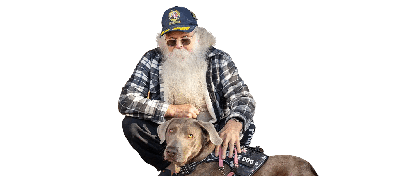 An older man with a long white beard, sunglasses, and a cap sits with a brown dog on a black background, wearing a plaid shirt and jacket from Joybound.