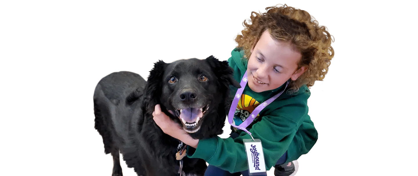 A curly-haired child in a green Joybound jacket hugs a black dog, isolated on a white background.