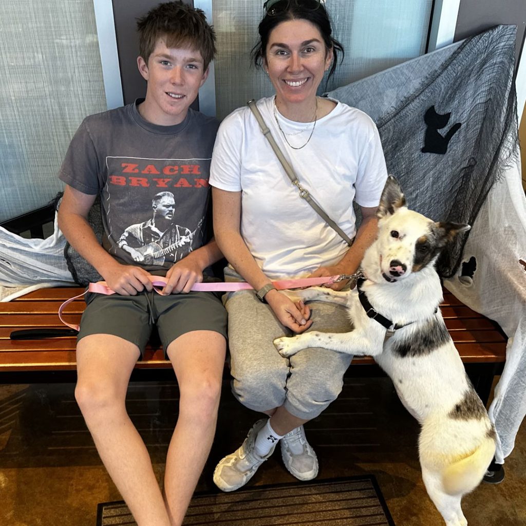 A woman and a boy sit on a wooden bench with a black-and-white dog on a leash in a pet adoption setting.